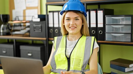 Photo for Young blonde woman architect smiling confident using laptop wearing hardhat at office - Royalty Free Image