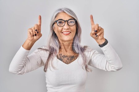 Téléchargez les photos : Femme d'âge moyen avec les cheveux gris debout sur fond blanc souriant étonné et surpris et pointant vers le haut avec les doigts et les bras levés. - en image libre de droit
