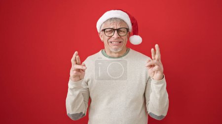 Photo for Middle age grey-haired man wearing christmas hat crossing fingers for lucky over isolated red background - Royalty Free Image