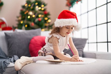 Foto de Adorable chica caucásica dibujando en el cuaderno sentado en el sofá por el árbol de Navidad en casa - Imagen libre de derechos