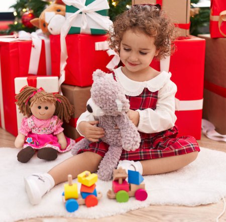 Photo for Adorable hispanic toddler playing with teddy bear sitting on floor by christmas gifts at home - Royalty Free Image