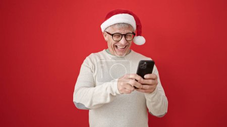 Téléchargez les photos : Homme moyen âge aux cheveux gris portant un chapeau de Noël en utilisant un smartphone sur fond rouge isolé - en image libre de droit