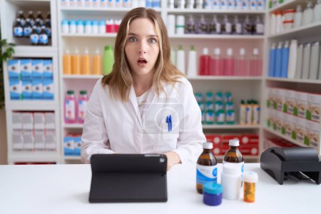 Téléchargez les photos : Jeune femme caucasienne travaillant à la pharmacie pharmacie utilisant comprimé peur et étonné avec la bouche ouverte pour la surprise, visage incrédule - en image libre de droit