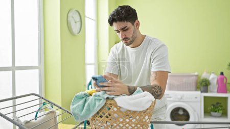 Photo for Young hispanic man using smartphone leaning on basket with clothes at laundry room - Royalty Free Image