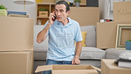 Photo for Middle age man unpacking cardboard box talking on smartphone at new home - Royalty Free Image