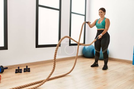 Photo for Young beautiful hispanic woman smiling confident using battle rope training at sport center - Royalty Free Image
