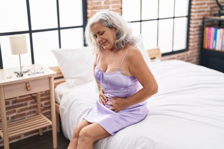 Téléchargez les photos : Femme aux cheveux gris d'âge moyen souffrant de douleurs menstruelles assise sur le lit dans la chambre - en image libre de droit
