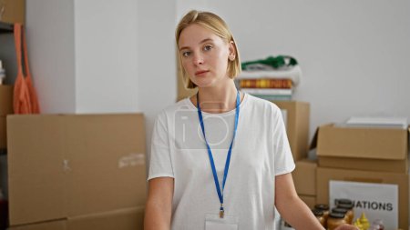 Téléchargez les photos : Jeune femme blonde bénévole debout avec un visage sérieux au centre de charité - en image libre de droit