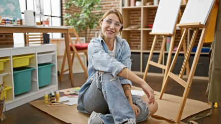 Téléchargez les photos : Jeune femme artiste souriant confiant assis sur le sol à l'atelier d'art - en image libre de droit