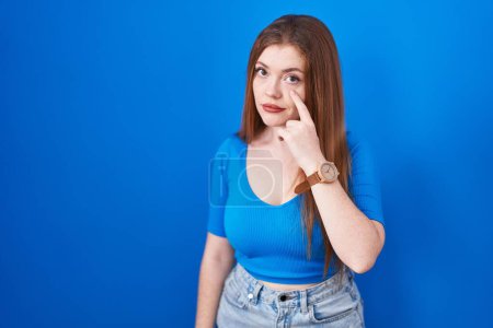 Photo for Redhead woman standing over blue background pointing to the eye watching you gesture, suspicious expression - Royalty Free Image
