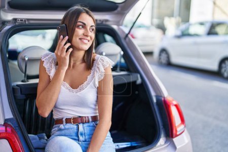Téléchargez les photos : Young beautiful hispanic woman talking on smartphone sitting on car trunk at street - en image libre de droit