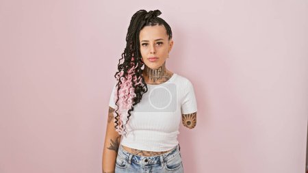 Téléchargez les photos : Cool femme amputée hispanique, debout sur fond rose isolé, exprimant sérieusement un mode de vie détendu avec un look tatoué sans bras. - en image libre de droit