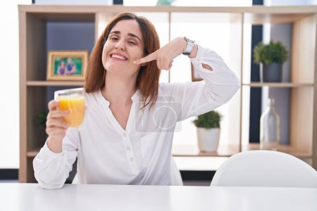 Téléchargez les photos : Femme brune buvant un verre de jus d'orange souriant et joyeux montrant et pointant du doigt les dents et la bouche. concept de santé dentaire. - en image libre de droit
