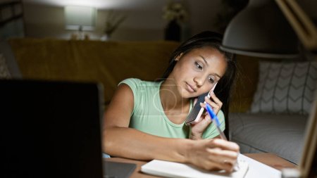 Photo for Young latin woman talking on smartphone using laptop taking notes at home - Royalty Free Image