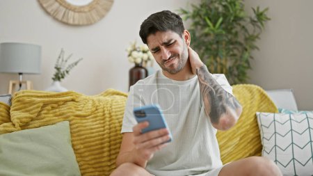 Téléchargez les photos : Jeune homme hispanique utilisant un smartphone assis sur un canapé souffrant de douleur cervicale à la maison - en image libre de droit