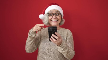 Téléchargez les photos : Femme du Moyen Âge aux cheveux gris utilisant un smartphone avec expression gagnante sur fond rouge isolé - en image libre de droit