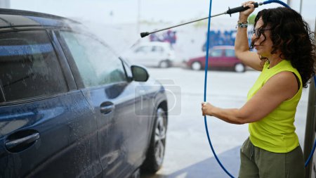 Photo for Middle age hispanic woman washing car with pressure washer at car wash station - Royalty Free Image