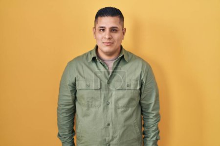Photo for Hispanic young man standing over yellow background relaxed with serious expression on face. simple and natural looking at the camera. - Royalty Free Image