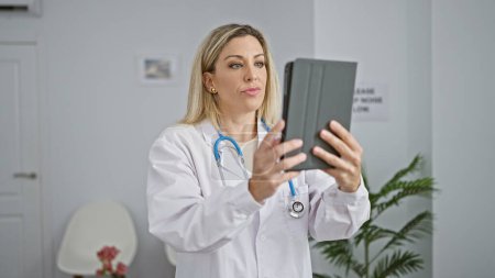 Téléchargez les photos : Jeune femme blonde médecin utilisant touchpad ith visage sérieux à la salle d'attente de la clinique - en image libre de droit