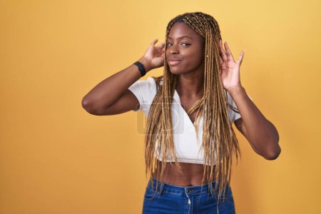 Téléchargez les photos : Femme afro-américaine aux cheveux tressés debout sur fond jaune essayant d'entendre les deux mains sur le geste de l'oreille, curieuse de commérages. problème auditif, sourd - en image libre de droit