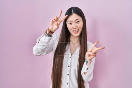 Téléchargez les photos : Jeune femme chinoise debout sur fond rose souriant avec la langue montrant les doigts des deux mains faisant signe de victoire. numéro deux. - en image libre de droit