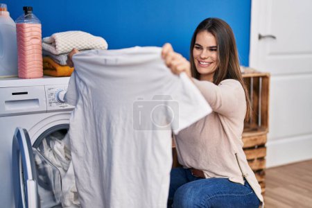 Foto de Joven hermosa mujer hispana sonriendo confiada lavando ropa en la lavandería - Imagen libre de derechos