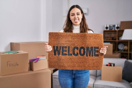 Photo for Young hispanic woman holding welcome doormat at new home sticking tongue out happy with funny expression. - Royalty Free Image