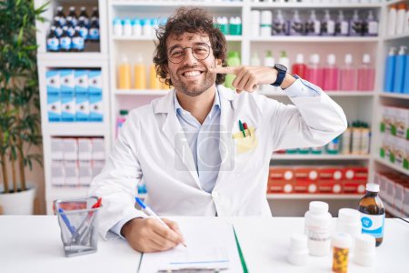 Téléchargez les photos : Jeune homme hispanique travaillant à la pharmacie pharmacie souriant gai montrant et pointant avec les doigts les dents et la bouche. concept de santé dentaire. - en image libre de droit