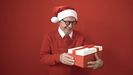 Téléchargez les photos : Senior portant chapeau de Noël déballage cadeau sur fond rouge isolé - en image libre de droit
