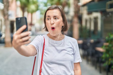 Foto de Mujer hispana de mediana edad haciendo videollamadas con smartphone asustada y sorprendida con la boca abierta para sorpresa, cara de incredulidad - Imagen libre de derechos