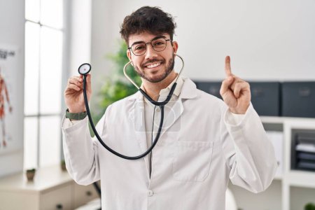 Photo for Hispanic man with beard wearing doctor uniform and stethoscope smiling with an idea or question pointing finger with happy face, number one - Royalty Free Image