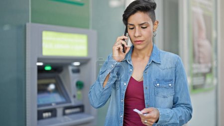 Téléchargez les photos : Jeune belle femme hispanique parlant sur smartphone tenant la carte de crédit au guichet de la banque - en image libre de droit
