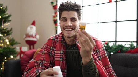 Photo for Young hispanic man holding pills bottle sitting on sofa by christmas tree at home - Royalty Free Image