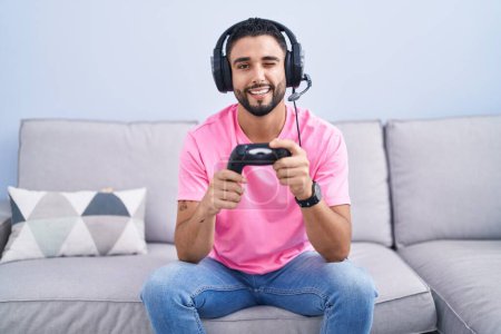 Téléchargez les photos : Jeune homme hispanique jouant contrôleur de tenue de jeu vidéo assis sur le canapé clin d'oeil en regardant la caméra avec une expression sexy, gai et heureux visage. - en image libre de droit