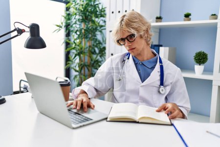 Foto de Young blond man doctor using laptop reading book at clinic - Imagen libre de derechos