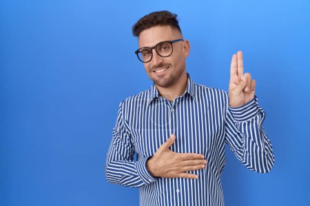 Foto de Hombre hispano con barba usando gafas sonriendo jurando con la mano en el pecho y los dedos hacia arriba, haciendo una promesa de lealtad juramento - Imagen libre de derechos