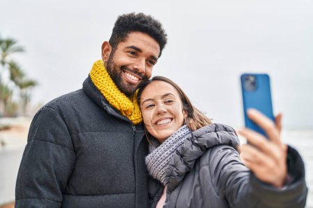 Téléchargez les photos : Homme et femme couple souriant confiant faire selfie par smartphone au bord de la mer - en image libre de droit