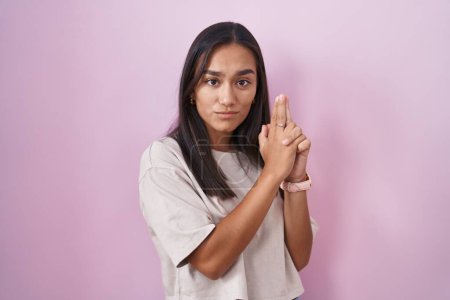 Téléchargez les photos : Young hispanic woman standing over pink background holding symbolic gun with hand gesture, playing killing shooting weapons, angry face - en image libre de droit