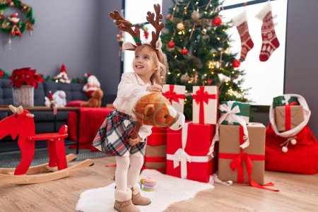 Photo for Adorable blonde girl playing with horse toy standing by christmas tree at home - Royalty Free Image