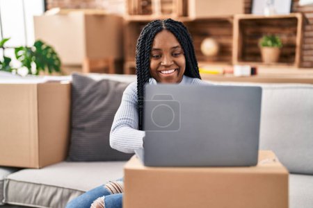 Téléchargez les photos : Femme afro-américaine utilisant un ordinateur portable assis sur le canapé à la nouvelle maison - en image libre de droit