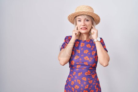 Foto de Mujer caucásica joven con vestido de flores y sombrero de verano que cubre las orejas con los dedos con expresión molesta por el ruido de la música fuerte. concepto de sordo. - Imagen libre de derechos