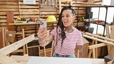 Photo for Smiling hispanic amputee woman, a talented carpenter, joyfully engages in a video call while indoors at her lively carpentry workshop - Royalty Free Image