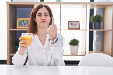 Photo for Brunette woman drinking glass of orange juice showing middle finger, impolite and rude fuck off expression - Royalty Free Image