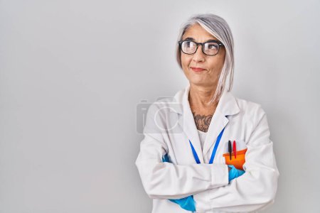 Foto de Mujer de mediana edad con el pelo gris vistiendo bata científica sonriendo mirando a un lado y mirando hacia otro pensando. - Imagen libre de derechos