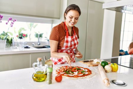 Téléchargez les photos : Young beautiful hispanic woman smiling confident putting onion on pizza at the kitchen - en image libre de droit