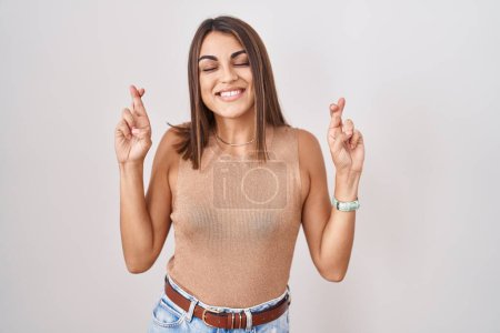 Téléchargez les photos : Young hispanic woman standing over white background gesturing finger crossed smiling with hope and eyes closed. luck and superstitious concept. - en image libre de droit