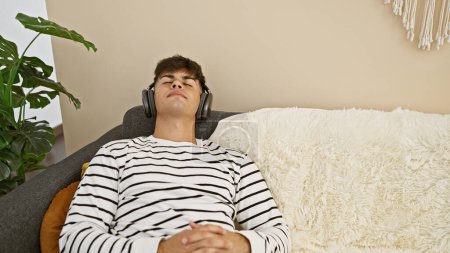 Foto de Alegre joven hispano en zona de auriculares escuchando música bombeada, disfrutando con confianza del divertido estilo de vida interior. él está reclinado en un sofá en casa, lleno de felicidad y sonriendo. - Imagen libre de derechos