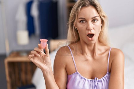 Foto de Mujer rubia joven sosteniendo la copa menstrual asustada y sorprendida con la boca abierta para sorpresa, cara de incredulidad - Imagen libre de derechos