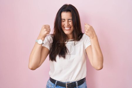Téléchargez les photos : Jeune femme brune debout sur fond rose excité pour le succès avec les bras levés et les yeux fermés célébrant la victoire en souriant. concept gagnant. - en image libre de droit