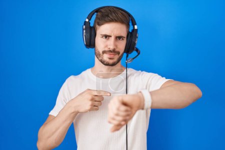 Téléchargez les photos : Homme hispanique avec la barbe à l'écoute de la musique portant des écouteurs à la hâte pointant vers regarder le temps, l'impatience, bouleversé et en colère pour retard de délai - en image libre de droit
